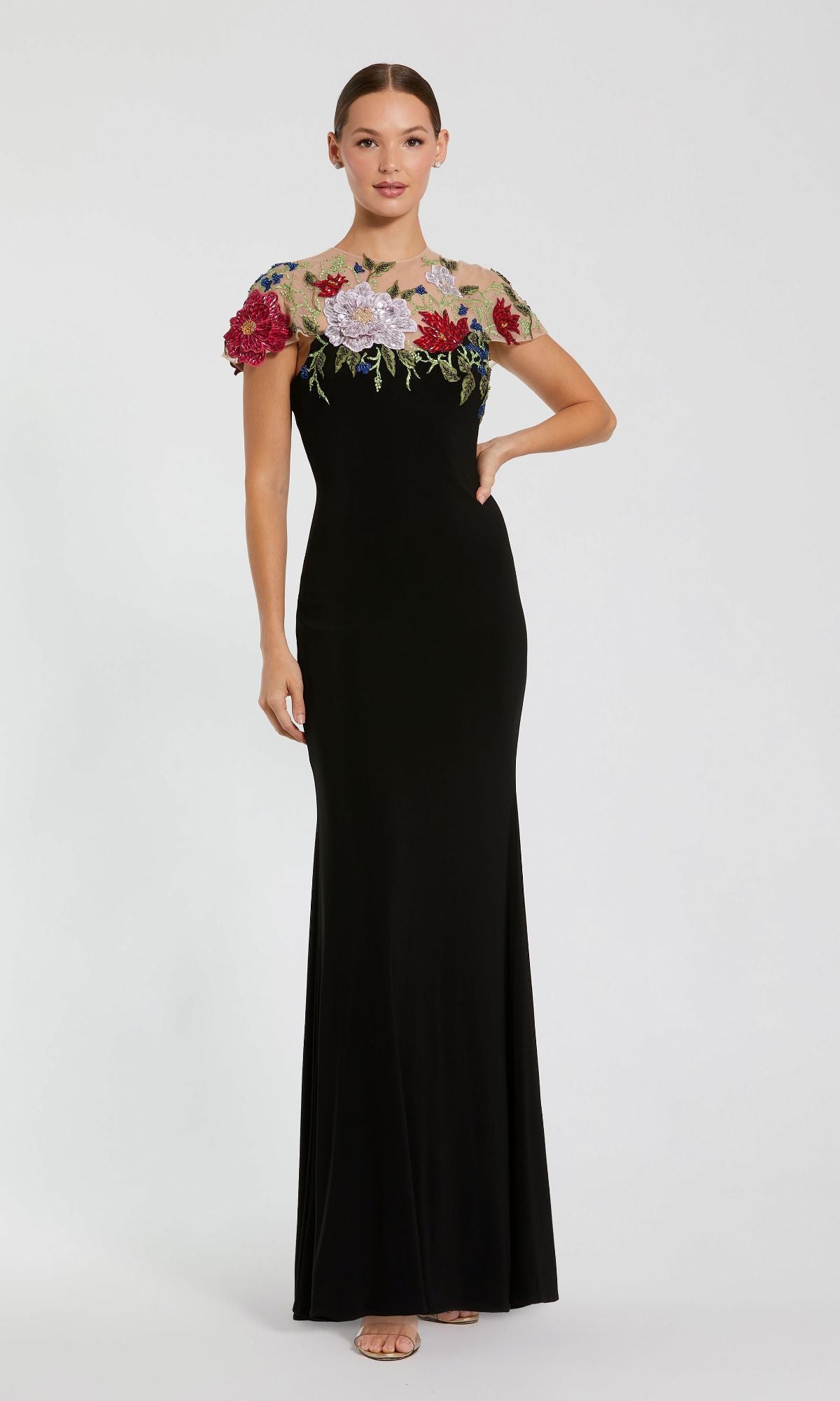 A woman wears the Mac Duggal 11817, a floral long black formal dress with short sheer sleeves and colorful floral embroidery on the bodice, standing against a plain background.